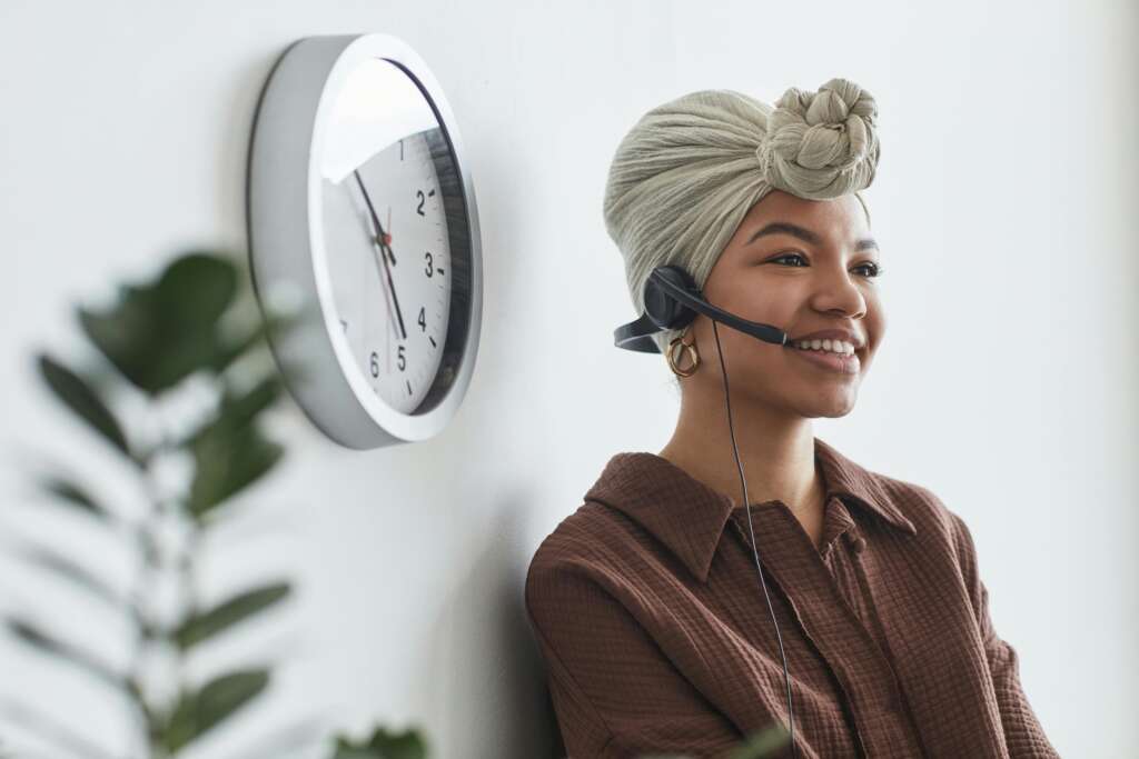 Cheerful woman in turban using headset in bright office space with clock on wall.